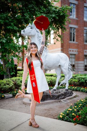 USC graduate in white dress and red Class of 2026 sash celebrates by white Trojan horse statue on campus.