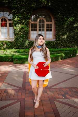 Smiling USC graduate in white dress holding red mortarboard cap on brick pathway with ivy-covered building backdrop.