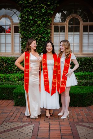 Three women in white dresses wearing red USC graduation stoles pose together outdoors on a brick path.