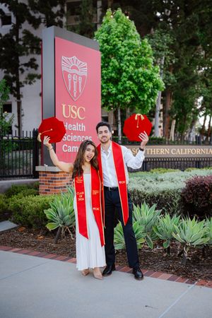 Two USC graduates in red stoles hold mortarboards in front of USC Health Sciences Campus sign.