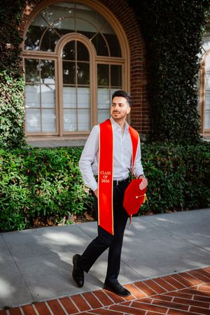 Young man in graduation attire wearing red USC Class of 2024 stole walks confidently on brick path near ivy-covered building.