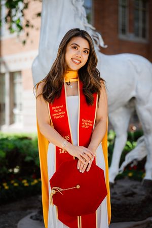USC graduate smiles in red and gold regalia with Doctor of Nurse Anesthesia Practice stole near Trojan horse statue.