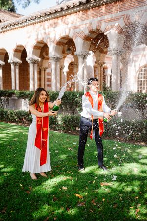 Two USC graduates in red sashes celebrate outdoors by spraying champagne near a historic arched building.