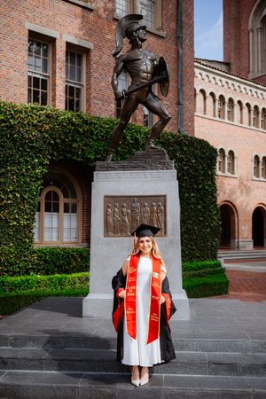 Graduate in cap and gown poses before Tommy Trojan statue at USC, wearing red and white honor stoles.