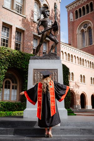 USC graduate in cap and gown stands before the Tommy Trojan statue on campus, wearing red and gold honor stoles.