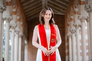 USC graduate wearing white dress and red graduation stole stands smiling in ornate columned hallway at Mudd Hall.