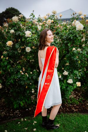 A person in a red USC graduation stole poses joyfully in front of blooming cream-colored roses in a garden.
