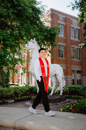 USC graduate in red sash walks proudly near white horse statue at brick university building surrounded by green trees.