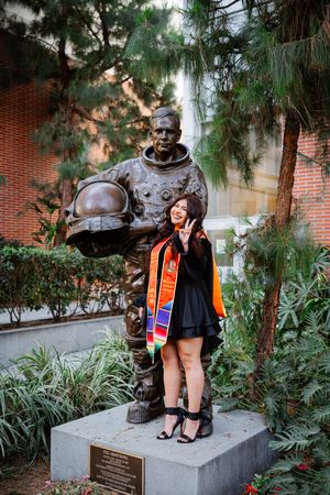 USC graduate in black gown and colorful stole poses with peace sign beside bronze astronaut statue outdoors at Viterbi.