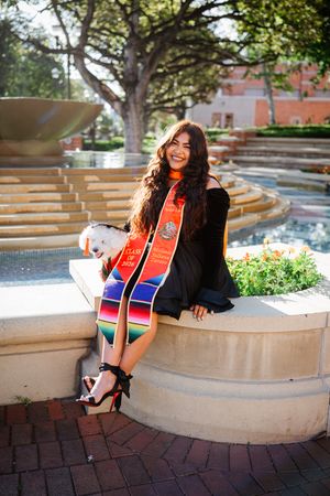 USC graduate in black gown with colorful sash sits by fountain, smiling, holding small white dog, wearing heels.