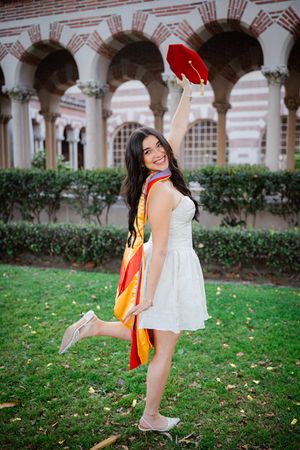 Young woman in white dress celebrating her USC graduation, holding red cap aloft outdoors near arched building.