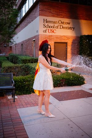 USC dental school graduate in white dress and red cap sprays champagne outside Herman Ostrow School of Dentistry.