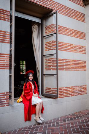 USC graduate in red cap and gown sitting in open window of brick building, smiling at camera on graduation day.