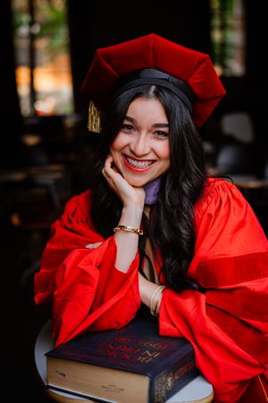 Smiling USC graduate in red cap and gown poses with books, celebrating academic achievement.