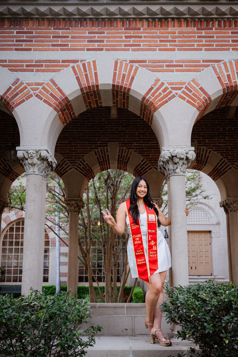 USC graduate in white dress and red sash poses under ornate brick archway with decorative columns at university campus.