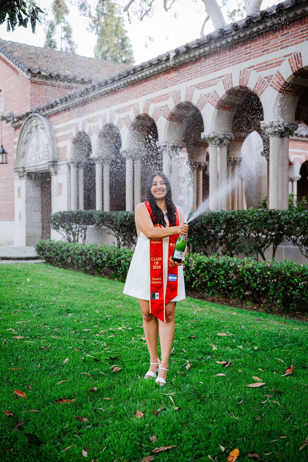 USC graduate in red and white regalia stands on grass lawn in front of Mudd Hall's ornate arched colonnade with brick architecture.