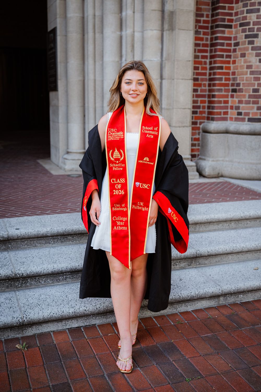 USC graduate in black cap and gown with red stole poses for graduation photos against brick building exterior.
