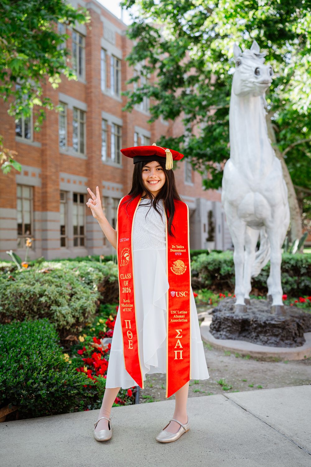 Graduate in red and white graduation stole poses with white lion statue on university campus grounds, USC.