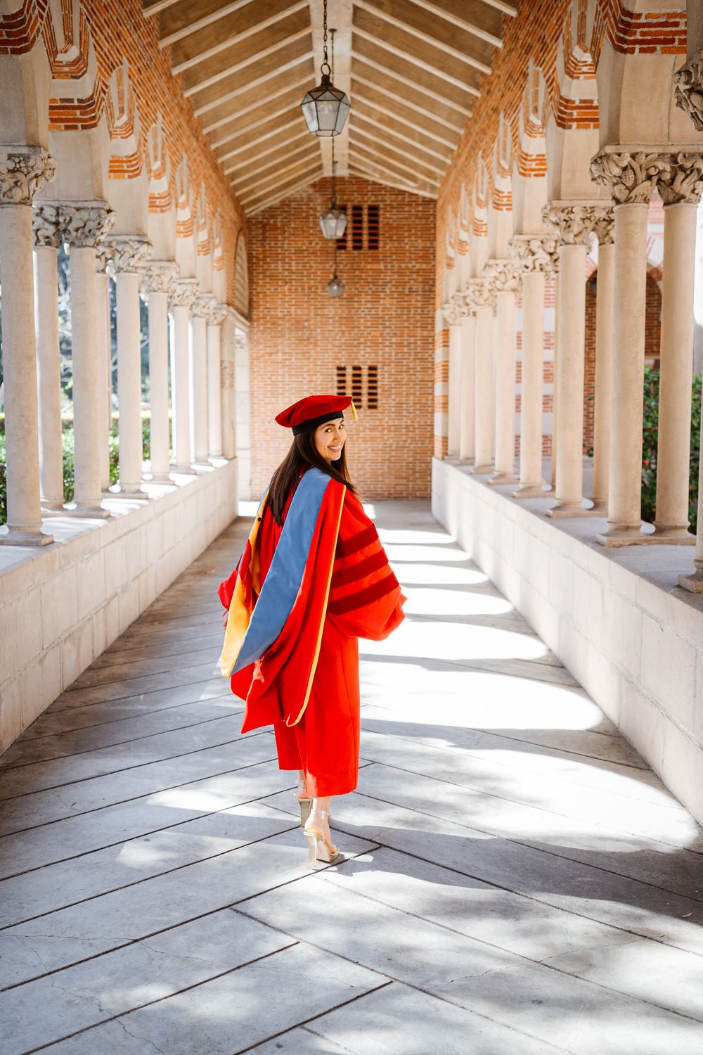 USC graduate in bright red academic regalia walks through ornate columned corridor with arched ceiling and brick walls.