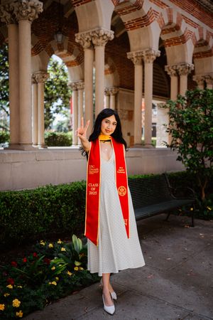 USC graduate in red stole and white dress flashing peace sign at Class of 2023 ceremony on campus.