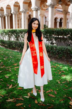 Young woman in white dress wearing red USC Class of 2025 graduation stole, posing outdoors on green lawn.