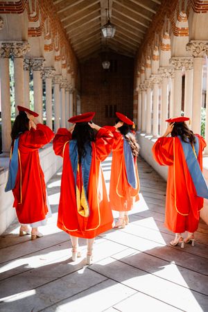Four PhD graduates in red doctoral robes and caps walk through an ornate university colonnade at Mudd Hall USC.