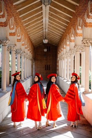 Four smiling PhD USC graduates in red doctoral robes hold hands in a sunlit university corridor.