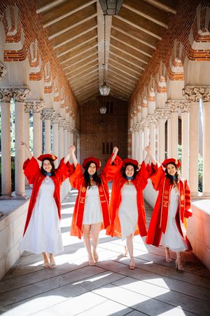 Four USC graduates in red robes and caps celebrate in an ornate university corridor, arms raised in joy.