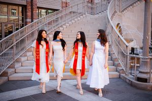 Four female USC graduates in white dresses with red sashes walk together on stone steps, celebrating their graduation day.
