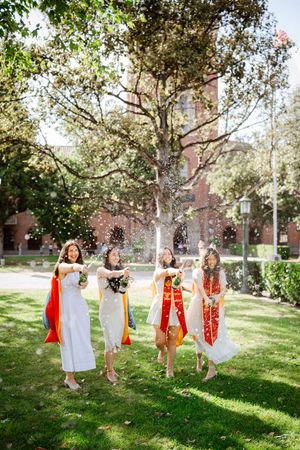 Four female USC graduates in white dresses and sashes celebrate outdoors with champagne on a sunny campus lawn at Alumni Park.