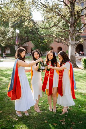 Four female USC graduates in white dresses with red sashes toast with champagne on a sunny campus lawn.