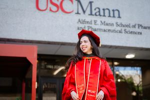 Graduate in red academic regalia and cap stands smiling outside USC Mann School of Pharmaceutical Sciences building.