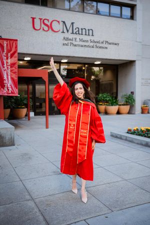 Graduate in red USC Mann School of Pharmacy regalia poses for celebratory photos outside campus building entrance.