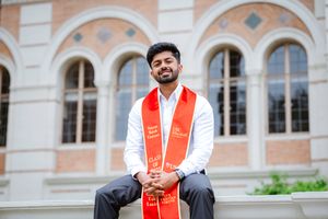 USC graduate in orange and white stole poses in front of ornate brick building with arched windows.