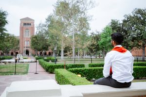 Students in white graduation gowns walk across the USC campus with brick buildings and green lawns in the background.