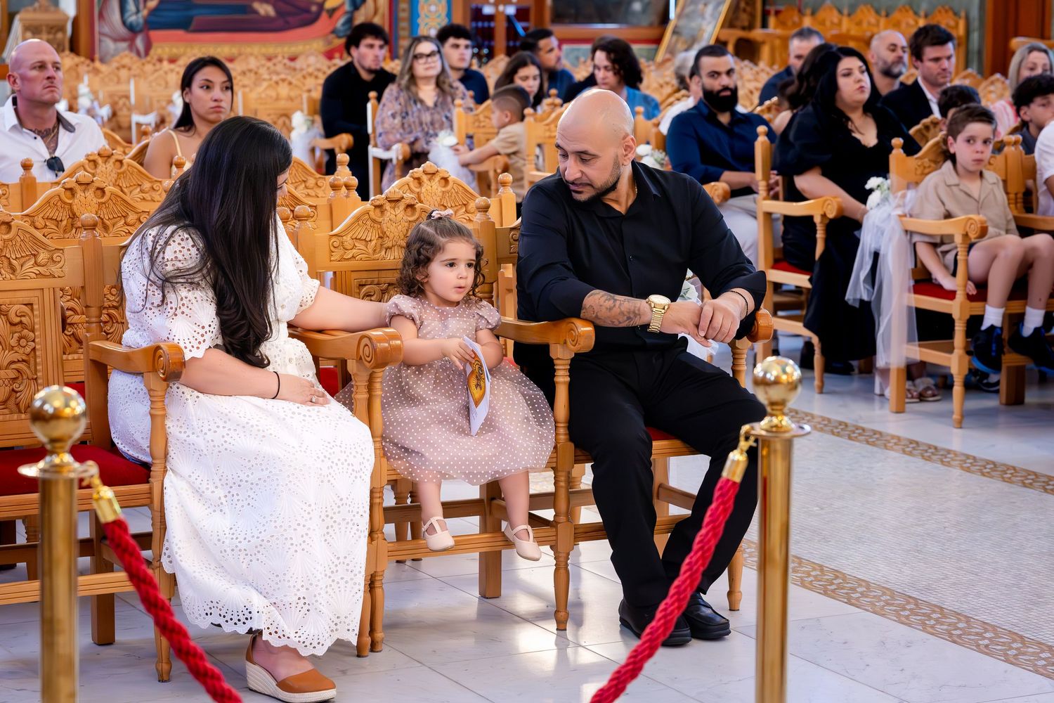 Family dressed formally seated in ornate church during a religious ceremony, with young girl in pink dress between parents.