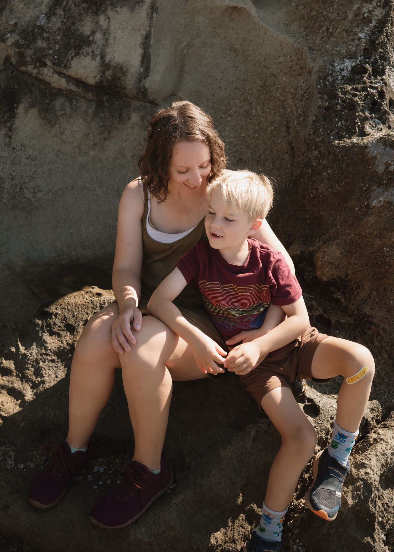 A woman and young blonde boy sit together on rocky terrain, smiling warmly in natural sunlight.