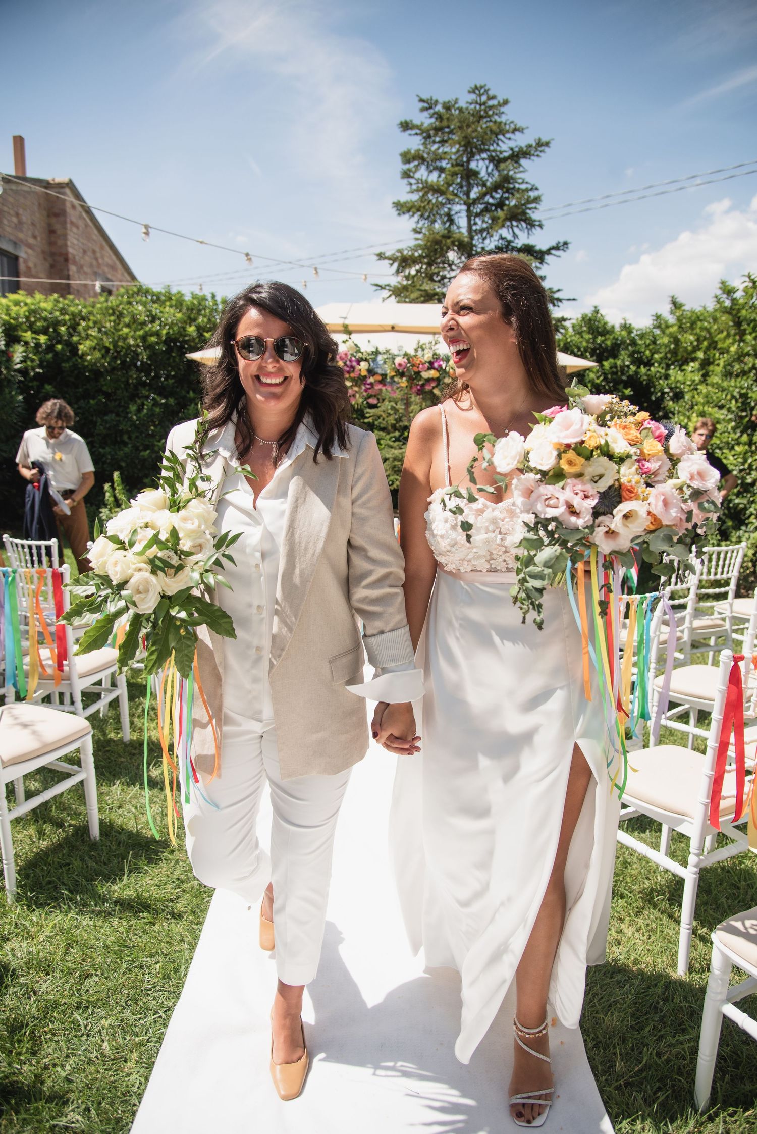 A couple in white and pink attire poses joyfully in a green outdoor setting on a cloudy day.