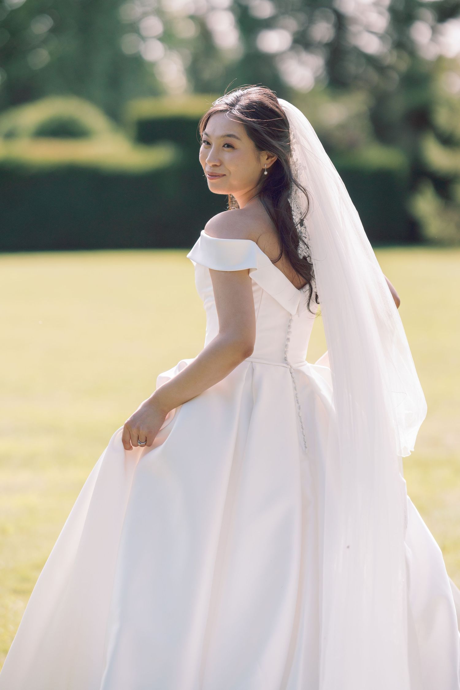 A bride in a flowing white wedding gown twirls gracefully in a sunlit meadow, her veil floating in the breeze at Kingston Maurward