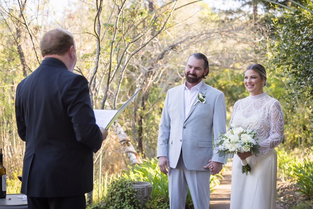 A group gathers outdoors for a wedding ceremony in a scenic wooded setting on a sunny day.