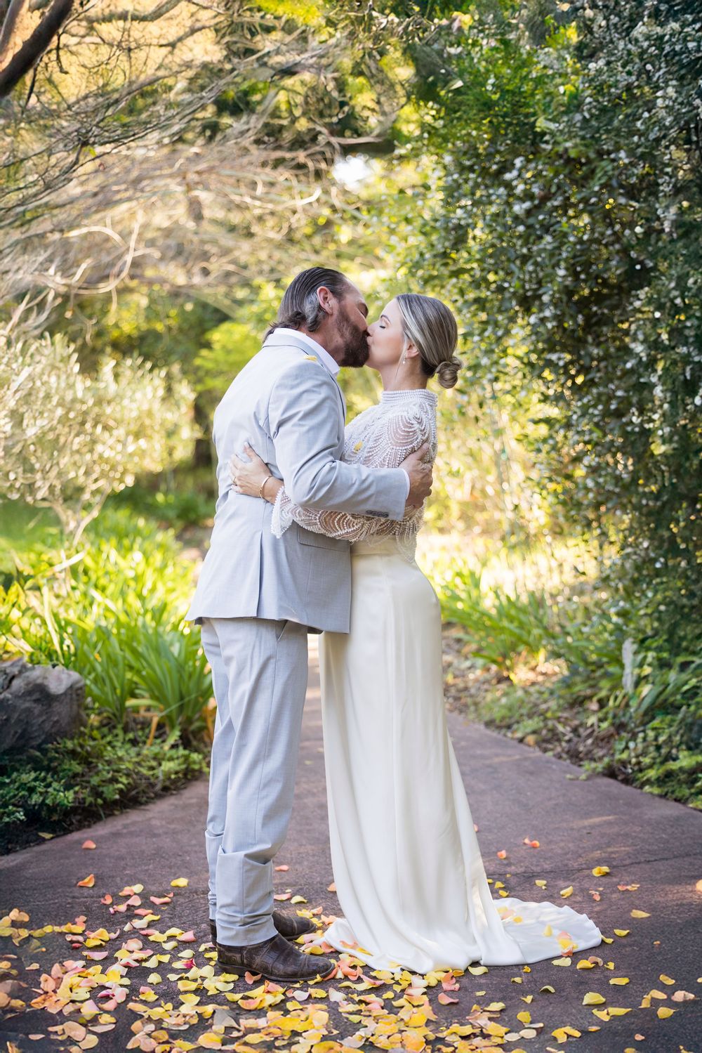A couple in white and grey attire dance together on a path with autumn leaves scattered around them.