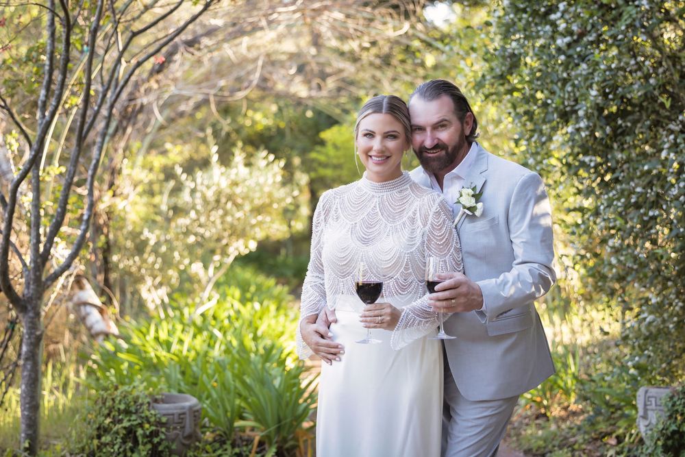 Couple in white attire sharing wine glasses together in a romantic outdoor garden setting at sunset.
