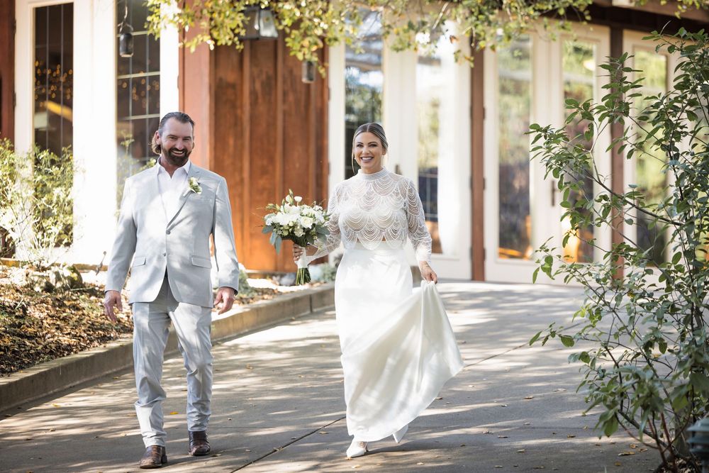 A couple walks together on a garden path, with the bride wearing a flowing white gown and the groom in a light suit.