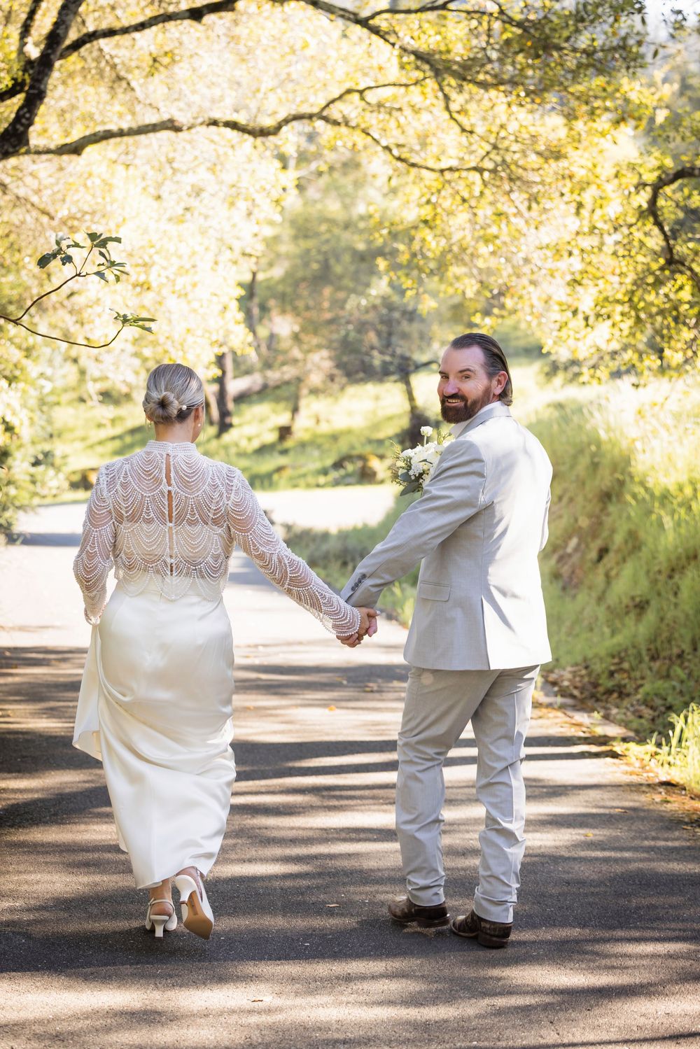 A couple in wedding attire walking hand in hand down a sunlit path surrounded by golden autumn foliage.