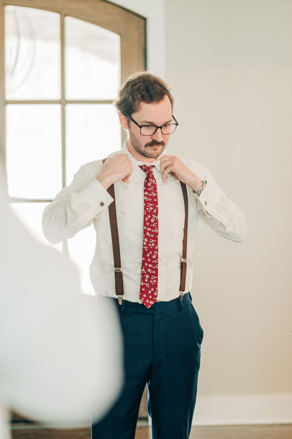 Professional portrait of person in vintage attire with suspenders and red tie against white wall in natural light.