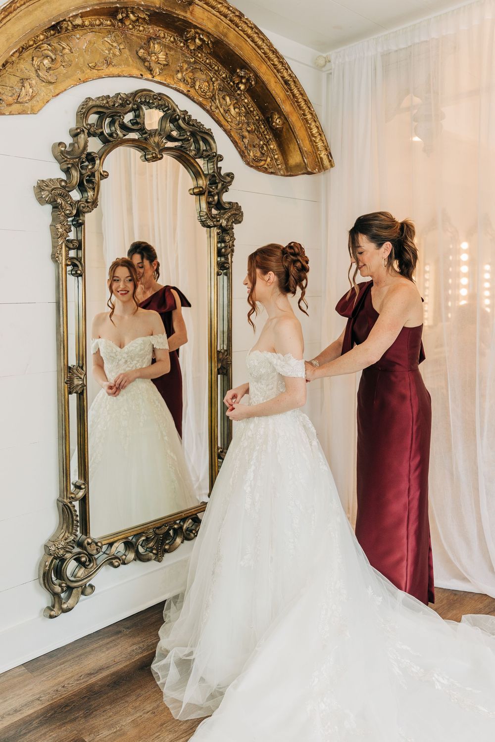 Bridal party in burgundy and white dresses gather around an ornate gold mirror in an elegant white room.