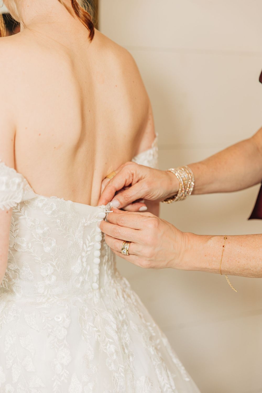 Hands lacing up the back of an elegant white wedding dress with delicate floral lace details.