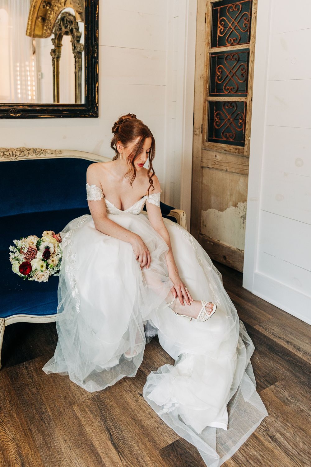 A bride in a flowing white wedding gown sits gracefully on rustic wooden flooring in an elegant indoor setting.