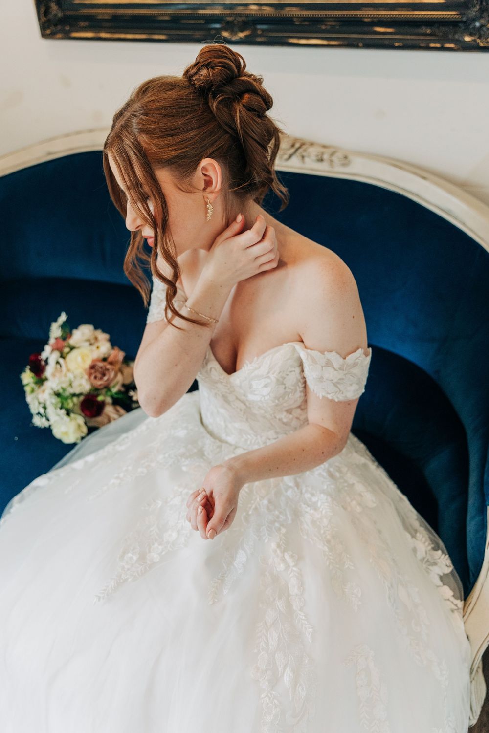 A bride in a white lace wedding dress sits gracefully on a blue velvet couch with a bouquet of flowers nearby.