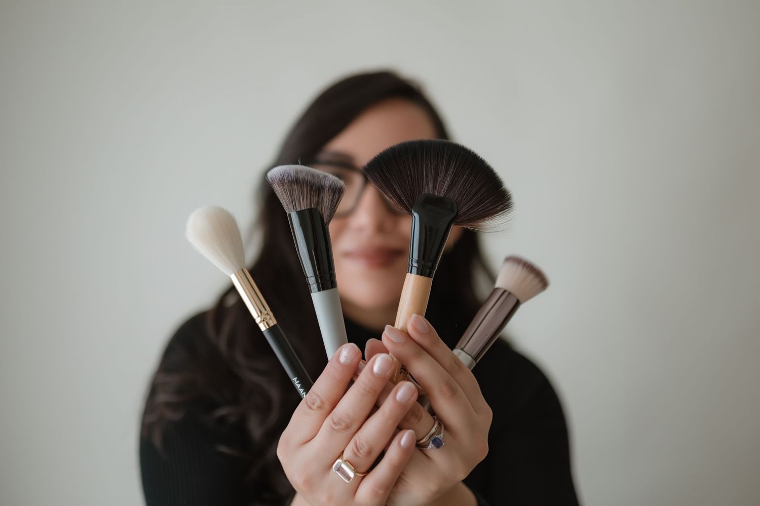 A person holding various makeup brushes and beauty tools against a neutral background.